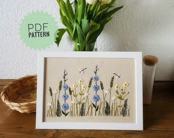 May include: A white frame with a beige background featuring an embroidered design of blue and yellow flowers, green leaves, and white dragonflies. The text "PDF PATTERN" is on a green circle in the top left corner of the image.
