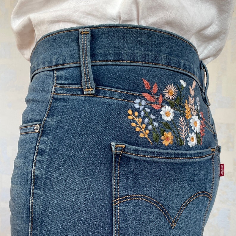 Boho-style floral embroidery design. White daisies and autumn wildflowers stitched above the back pocket of blue jeans. Hand-embroidered botanical elements creating a unique look for denim apparel.