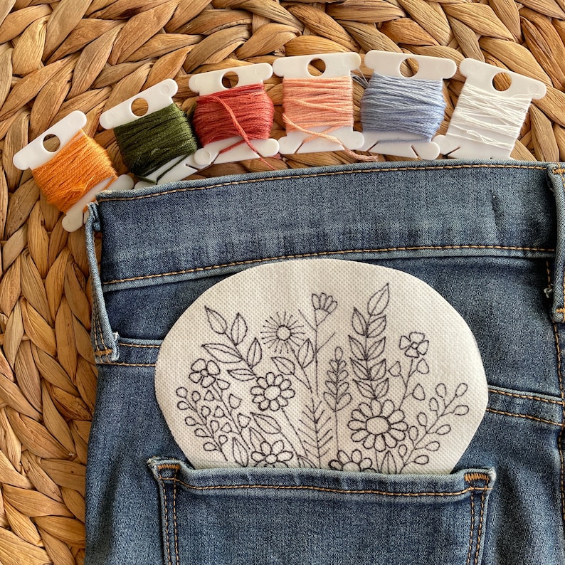 Wildflower embroidery PDF pattern color palette. Water soluble stabilizer with a floral outline tucked into a denim pocket, paired with six embroidery floss bobbins in mustard, olive green, terracotta, peach, blue, and white.