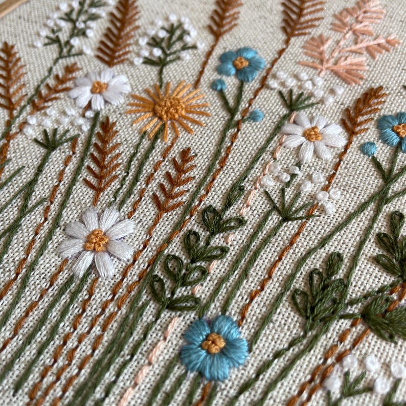 K&ouml;nnte beinhalten: Ein Stickrahmen mit einem botanischen Motiv auf beigem Stoff. Das Muster zeigt wei&szlig;e G&auml;nsebl&uuml;mchen mit gelben Mitten, blaue Blumen sowie verschiedene gr&uuml;ne und braune St&auml;ngel, Farne und Gr&auml;ser, die mit strukturiertem Garn gestickt sind.