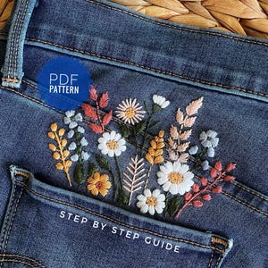 A pair of blue denim jeans featuring a detailed floral embroidery design on the back pocket. The embroidery includes white daisies, yellow wheat stalks, and pink sprigs. Text on the image reads PDF PATTERN and STEP BY STEP GUIDE.