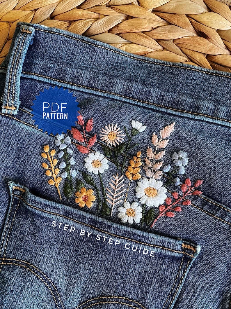 A pair of blue denim jeans featuring a detailed floral embroidery design on the back pocket. The embroidery includes white daisies, yellow wheat stalks, and pink sprigs. Text on the image reads PDF PATTERN and STEP BY STEP GUIDE.