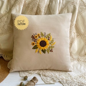 May include: A beige pillow with a sunflower embroidery design. The sunflower is surrounded by green leaves and brown stems. The pillow has the text "PDF PATTERN" on it.