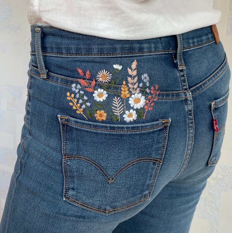A person wearing blue denim jeans, showcasing the hand-embroidered floral pocket from behind. Wildflowers embroidery on the pocket in warm tones. Digital PDF download for hand embroidery on jeans or shorts.