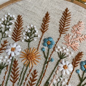 K&ouml;nnte beinhalten: Ein Stickrahmen mit beigefarbenem Leinenstoff und handgestickten botanischen Motiven. Das Muster zeigt wei&szlig;e G&auml;nsebl&uuml;mchen, gelbe Sonnenblumen, blaue Blumen sowie verschiedene weizenartige Halme und Gr&auml;ser in Braun- und Pfirsicht&ouml;nen.
