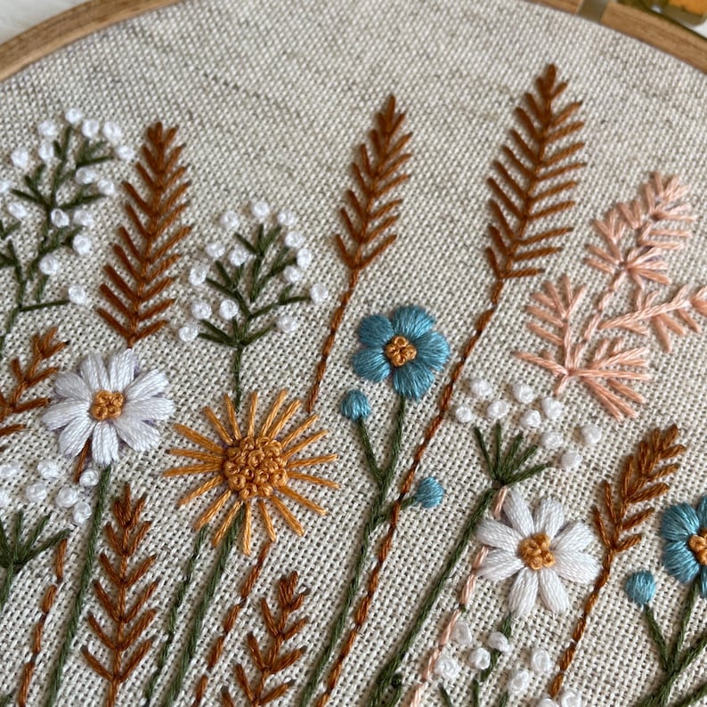 K&ouml;nnte beinhalten: Ein Stickrahmen mit beigefarbenem Leinenstoff und handgestickten botanischen Motiven. Das Muster zeigt wei&szlig;e G&auml;nsebl&uuml;mchen, gelbe Sonnenblumen, blaue Blumen sowie verschiedene weizenartige Halme und Gr&auml;ser in Braun- und Pfirsicht&ouml;nen.