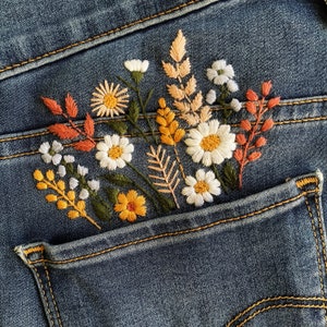 Boho floral embroidery design on back denim pocket. Detailed stitching of meadow flowers in white, mustard yellow, and terracotta orange. Seasonal autumn flower pattern for personalizing clothing.