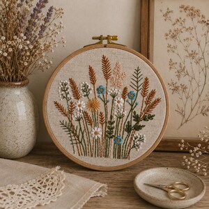 K&ouml;nnte beinhalten: Ein runder Stickrahmen aus Holz mit einem hellbeigen Stoff, der ein handgesticktes Motiv aus verschiedenen Wildblumen und Gr&auml;sern in Blau-, Wei&szlig;-, Gelb-, Gr&uuml;n- und Braunt&ouml;nen zeigt, inklusive einer Messingschraube am oberen Rand.