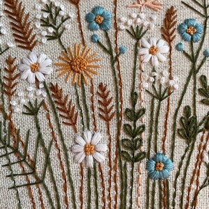 K&ouml;nnte beinhalten: Eine Stickerei auf beigefarbenem Leinenstoff, die ein Feld mit Wildblumen darstellt. Das Design umfasst wei&szlig;e G&auml;nsebl&uuml;mchen, gelbe sonnenf&ouml;rmige Blumen und kleine blaue Bl&uuml;ten, die durch gr&uuml;ne St&auml;ngel und braune weizenartige Halme verbunden sind.