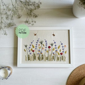 May include: A white frame with a beige background featuring an embroidered design of a field of wildflowers in shades of purple, pink, yellow, and green. Two small yellow bees are embroidered above the flowers. The text "PDF PATTERN" is on a green starburst shape in the top left corner of the image.