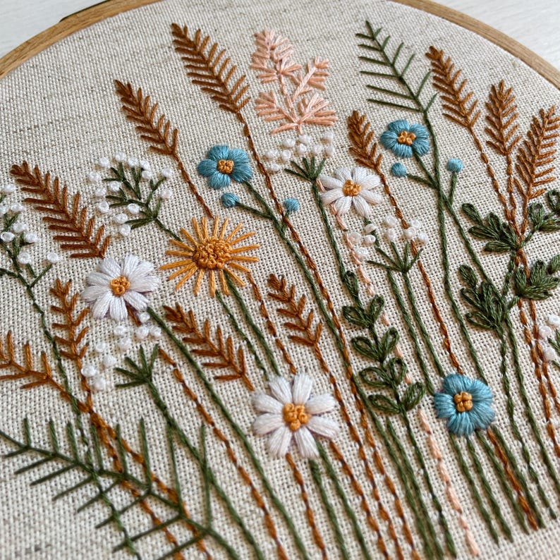K&ouml;nnte beinhalten: Ein runder Stickrahmen aus Holz mit einem hellbeigen Stoff, der ein handgesticktes botanisches Motiv zeigt. Die Stickerei besteht aus verschiedenen Wildblumen, Gr&auml;sern und Weizen&auml;hren in den Farben Blau, Wei&szlig;, Gelb und Braun.