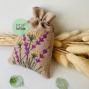 Puede incluir: Una pequeña bolsa de arpillera con cordón de ajuste, bordada con un diseño de flores de lavanda. La bolsa está llena de lavanda seca y tiene un estilo rústico y campestre.