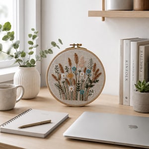 K&ouml;nnte beinhalten: Ein runder Stickrahmen aus Holz mit einem beigen Stoffeinsatz, der handgestickte botanische Motive zeigt. Die Stickerei besteht aus verschiedenen Wildblumen, Gr&auml;sern und Weizen&auml;hren in den Farben Wei&szlig;, Blau, Gelb und Braun auf hellem Grund.