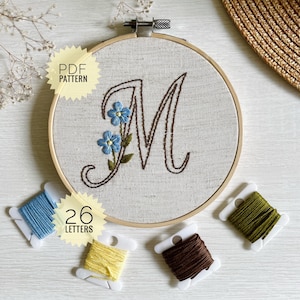 May include: An embroidered letter "M" in brown thread, decorated with blue flowers, within a wooden hoop. The text "PDF PATTERN" and "26 LETTERS" are present. Spools of blue, yellow, brown, and green embroidery floss are arranged around the hoop.