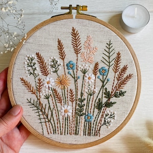 K&ouml;nnte beinhalten: Ein runder Stickrahmen aus Holz mit einem hellbeigen Stoff, der ein handgesticktes Motiv aus verschiedenen Wildblumen und Gr&auml;sern in Wei&szlig;, Braun, Gr&uuml;n, Gelb und Hellblau zeigt und mit einer metallenen Spannschraube versehen ist.