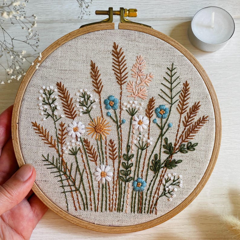 K&ouml;nnte beinhalten: Ein runder Stickrahmen aus Holz mit einem hellbeigen Stoff, der ein handgesticktes Motiv aus verschiedenen Wildblumen und Gr&auml;sern in Wei&szlig;, Braun, Gr&uuml;n, Gelb und Hellblau zeigt und mit einer metallenen Spannschraube versehen ist.