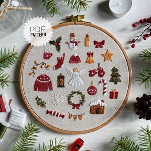 Puede incluir: Un aro de bordado blanco con un fondo de tela beige que presenta una variedad de diseños de bordado con temática navideña. Los diseños incluyen un muñeco de nieve, un árbol de Navidad, una media, una campana, un bastón de caramelo, una casa de jengibre, una corona, un copo de nieve, una linterna, una estrella, un regalo, una manopla, una taza de chocolate caliente, una cadena de luces, un adorno navideño y un ángel. El aro está rodeado de ramas de pino verdes y una vela blanca.