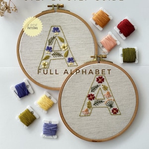 May include: Two wooden embroidery hoops containing fabric with stitched letter A designs featuring floral and leaf motifs. The text reads STEP BY STEP GUIDE, 2 PDF PATTERNS, and FULL ALPHABET. Several colourful embroidery thread bobbins are arranged nearby.