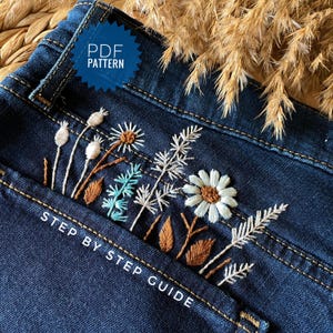 Peut inclure: Tissu en jean bleu foncé avec une broderie complexe de marguerites blanches, de feuilles brunes et de brins bleu clair. Un badge bleu en forme d'étoile affiche le texte PDF PATTERN, et le texte blanc STEP BY STEP GUIDE est imprimé en bas.