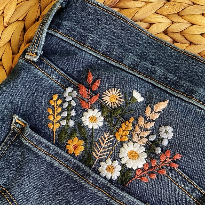 A close-up of a pair of blue denim jeans with a floral embroidery design. The embroidery features a variety of flowers in shades of orange, yellow, white, and pink.