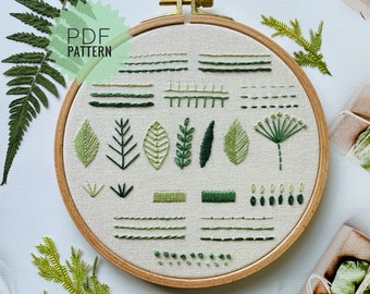 Puede incluir: Primer plano de un aro de bordado circular con un fondo de tela blanca. La tela está bordada con varios diseños de hojas y tallos verdes, incluyendo un helecho, una flor y una variedad de hojas. El aro es de madera y tiene un anillo de metal dorado.