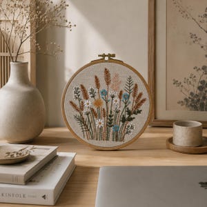 K&ouml;nnte beinhalten: Ein runder h&ouml;lzerner Stickrahmen mit einem Stoffeinsatz, der handgestickte botanische Motive zeigt. Die Stickerei besteht aus verschiedenen Wildblumen, Gr&auml;sern und St&auml;ngeln in Blau-, Wei&szlig;-, Gelb- und Braunt&ouml;nen auf einem hellbeigen Stoffhintergrund.