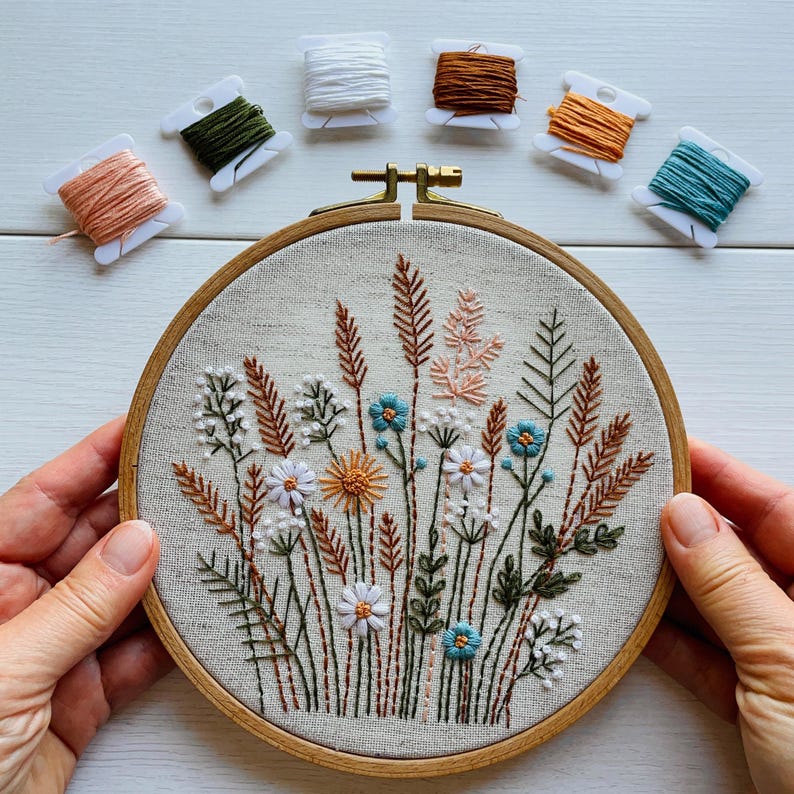 K&ouml;nnte beinhalten: Ein runder h&ouml;lzerner Stickrahmen mit beigem Stoff, der ein handgesticktes botanisches Design aus Wildblumen und Gr&auml;sern in Blau-, Wei&szlig;-, Orange- und Gr&uuml;nt&ouml;nen zeigt, umgeben von sechs kleinen Kunststoffspulen mit passendem Stickgarn.