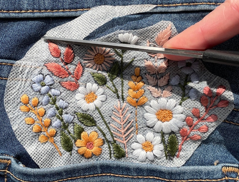 Trimming excess water soluble stabilizer from a wildflower embroidery design on a denim pocket. Hands using scissors to cut away the stabilizer from the finished hand-stitched floral bouquet. Visual guide for a DIY clothing upcycle PDF pattern.