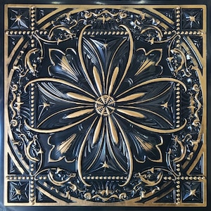 May include: A square, decorative panel in black and gold. The design features a central floral motif surrounded by ornate, embossed detailing. The gold highlights the raised patterns of leaves, stars, and geometric shapes, creating a striking contrast against the black.