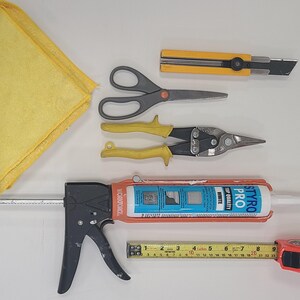 May include: A collection of construction tools including a yellow cloth, scissors, metal shears, a caulk gun, a tape measure, and a utility knife. The tape measure shows measurements in centimetres.
