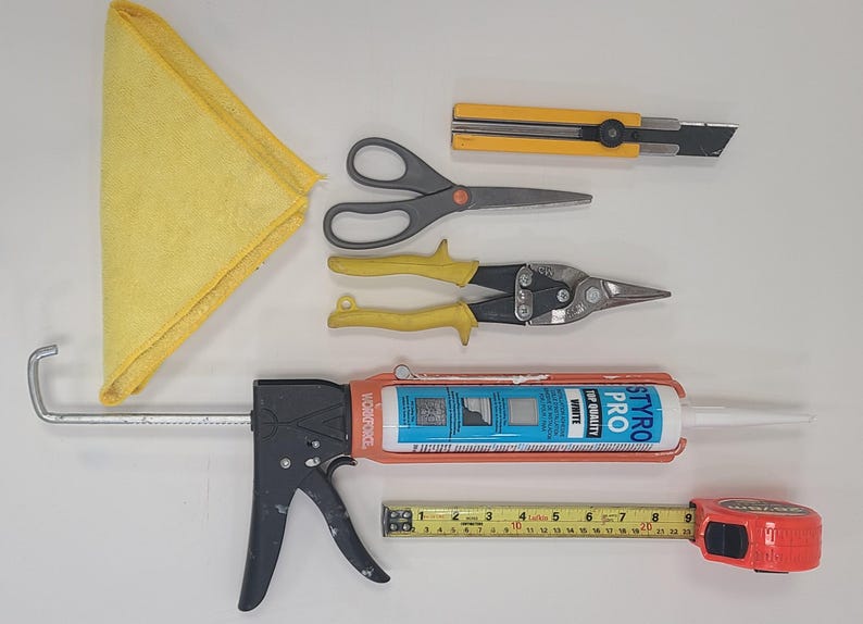 May include: A collection of construction tools including a yellow cloth, scissors, metal shears, a caulk gun, a tape measure, and a utility knife. The tape measure shows measurements in centimetres.