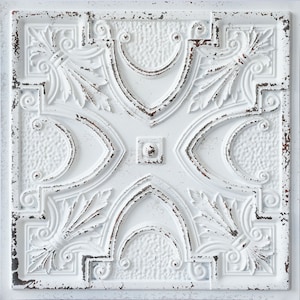 Op de afbeelding: Vierkante, witte decoratieve plafondtegel met een verweerde, antieke afwerking. De tegel heeft een herhalend patroon van bloem- en geometrische vormen, met een centraal vierkant en gebogen randen. Het ontwerp omvat verhoogde reliëfdetails en een getextureerd oppervlak.