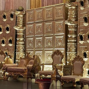 May include: An opulent gold wedding stage set. The backdrop is a large panel with a repeating pattern. The stage is furnished with gold chairs, a sofa, and decorative pillars. The design incorporates detailed carvings and architectural features.