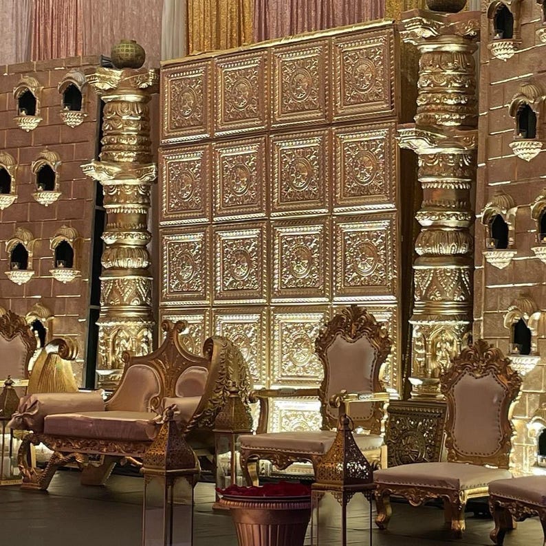 May include: An opulent gold wedding stage set. The backdrop is a large panel with a repeating pattern. The stage is furnished with gold chairs, a sofa, and decorative pillars. The design incorporates detailed carvings and architectural features.