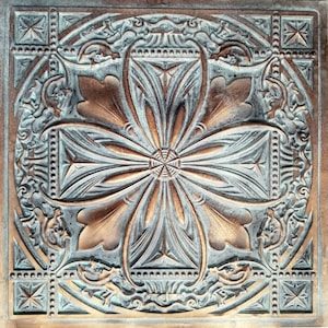 May include: A square, copper-coloured tile with a raised, intricate design. The design features a central flower with four petals, surrounded by a decorative border.