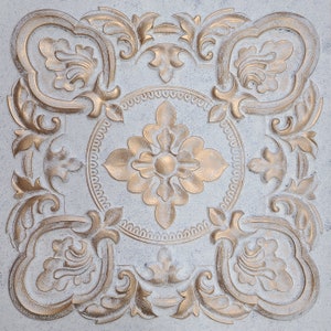 May include: A white square tile with a gold embossed design. The design features a central flower with four petals and a circle of smaller circles around it. The corners of the tile have a more intricate design with swirls and leaves.