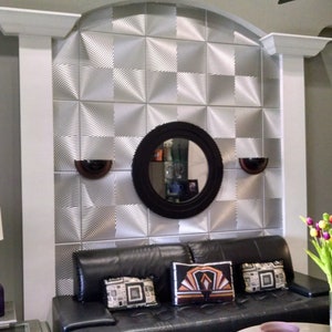 May include: A living room with a gray wall, white columns, and a black leather sofa. The wall behind the sofa is covered in silver metallic tiles with a geometric pattern. A round mirror with a black frame hangs on the wall above the sofa. There are two wall sconces on either side of the mirror. A vase with colorful flowers sits on a table in front of the sofa.