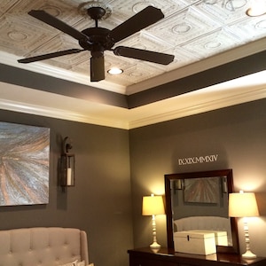 May include: A bedroom with a white ceiling with decorative tile panels, a ceiling fan, a brown dresser with six drawers, two lamps, a mirror with the text "IX.XIX.MMXIV", and a bed with a beige headboard and pillows.