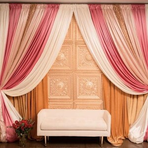 May include: A decorative backdrop with layered curtains in pink, cream, and gold, and a cream-colored bench. The backdrop includes a textured wall panel and a vase of red roses.