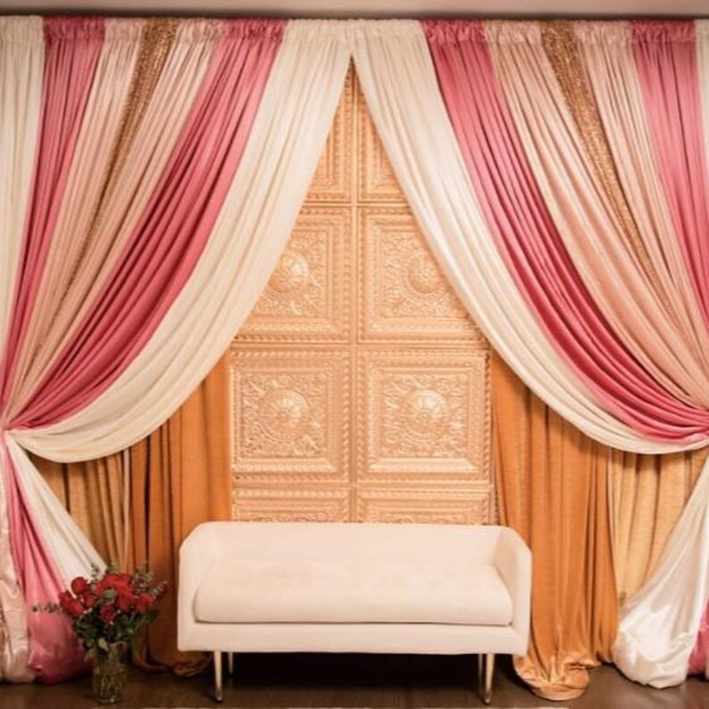 May include: A decorative backdrop with layered curtains in pink, cream, and gold, and a cream-colored bench. The backdrop includes a textured wall panel and a vase of red roses.