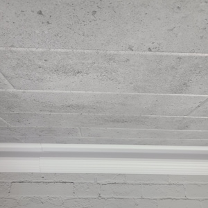 May include: A close-up view of a light gray, textured wall with horizontal lines and a white trim at the bottom. The wall has a rough, stone-like appearance, with subtle variations in color and texture.