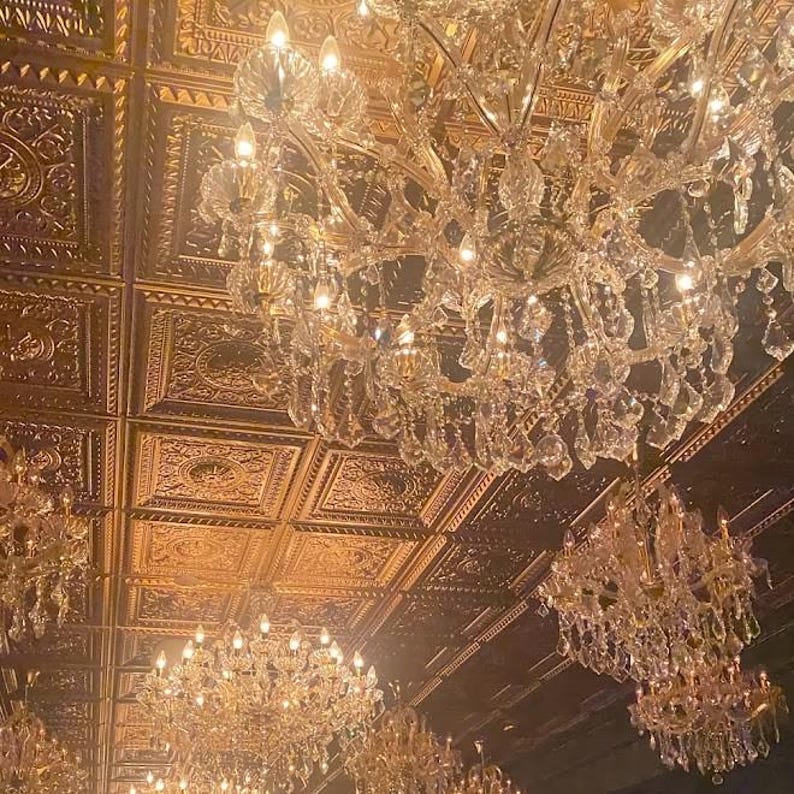 May include: Several crystal chandeliers hang from a decorative, gold-toned ceiling. The chandeliers are lit, creating a warm ambience. The ceiling features a repeating square pattern with embossed detailing. The overall style is luxurious and elegant.