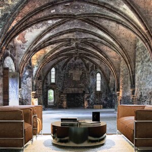 May include: Two brown leather sofas with metal frames sit facing each other in a large, vaulted room with stone walls. A round coffee table with a glass top sits between the sofas. The room has a high, arched ceiling and large windows.