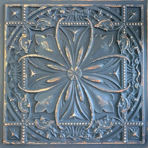 May include: A decorative ceiling tile with a distressed, dark blue finish and a gold floral design. The tile has a raised, ornate pattern with a central flower motif.