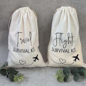 May include: Two white cotton drawstring bags with black text. The bag on the left says "Travel Survival Kit" and the bag on the right says "Flight Survival Kit". Both bags have a small airplane and heart design.