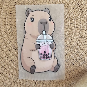 Könnte beinhalten: Cartoon-Illustration eines Capybaras, das ein Bubble Tea hält. Das Capybara ist hellbraun mit rosa Wangen und dunklen Augen. Der Bubble Tea ist rosa mit schwarzen Tapiokaperlen in einem transparenten Plastikbecher.
