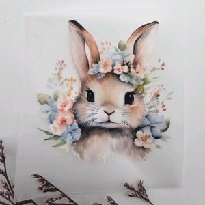 Könnte beinhalten: Aquarell-Illustration eines Hasen mit Blumenkranz. Der Hase hat braunes Fell, lange Ohren und einen lieben Gesichtsausdruck. Der Kranz zeigt blaue und rosa Blumen mit grünen Blättern, vor weißem Hintergrund.
