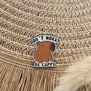 Könnte beinhalten: Ein braun-weißer Emaille-Pin mit einem Cartoon-Capybara und dem Text "DON'T WORRY BE CAPPY". Der Pin ist vor einem Hintergrund aus gewebtem Stroh platziert.
