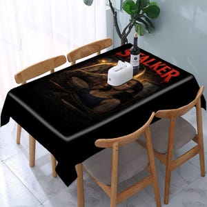 May include: Black rectangular tablecloth with a white border, featuring a graphic design with the word "STALKER" in red. The design includes a person and a bowl. The tablecloth is on a wooden table with wooden chairs.