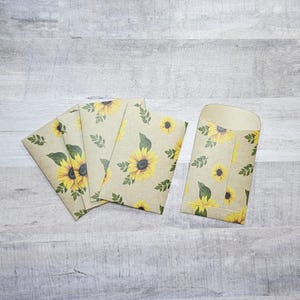 May include: Set of five kraft paper envelopes with a sunflower pattern. The envelopes are brown with a white background and feature yellow sunflowers with green leaves.